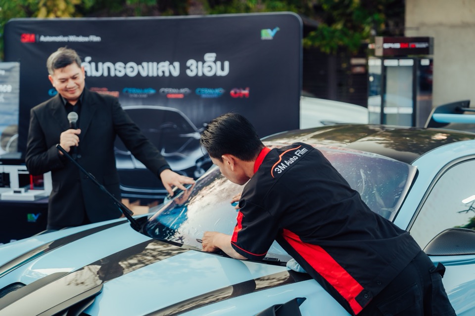 วิทยากรถือไมค์ยืนข้างช่างที่สาธิตการติดฟิล์มบนฝากระโปรงรถ หลังฉากมีป้ายฟิล์มกรองแสง 3M